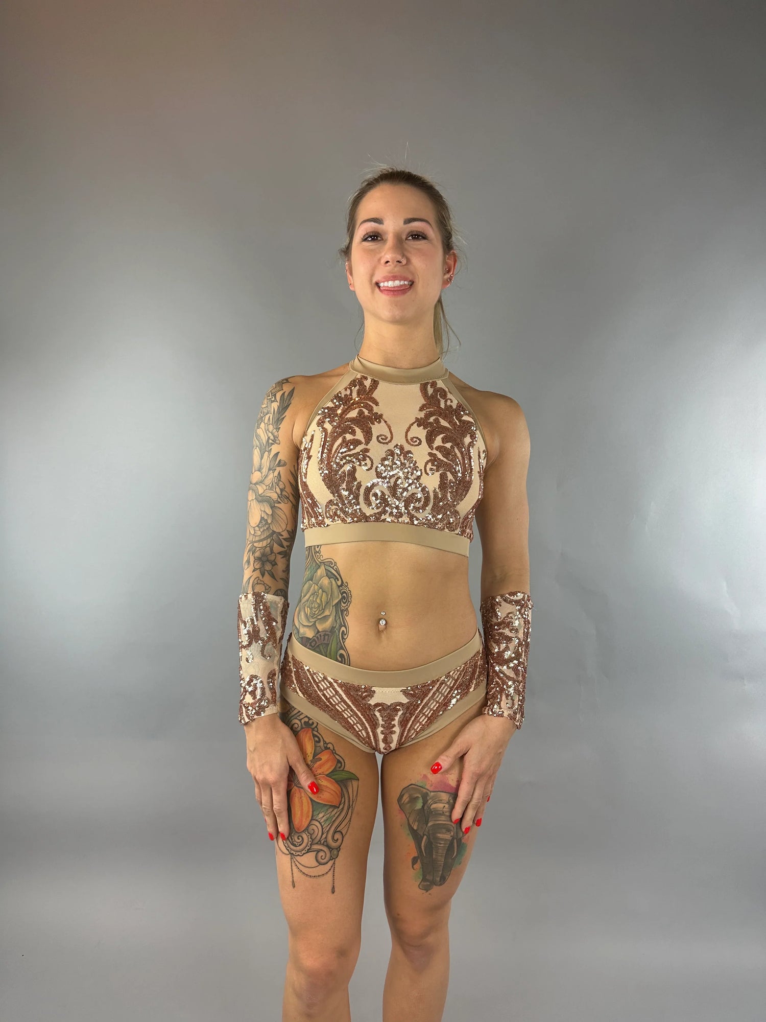 Woman in a patterned beige and brown bikini, smiling, with visible arm and leg tattoos, against a gray background.