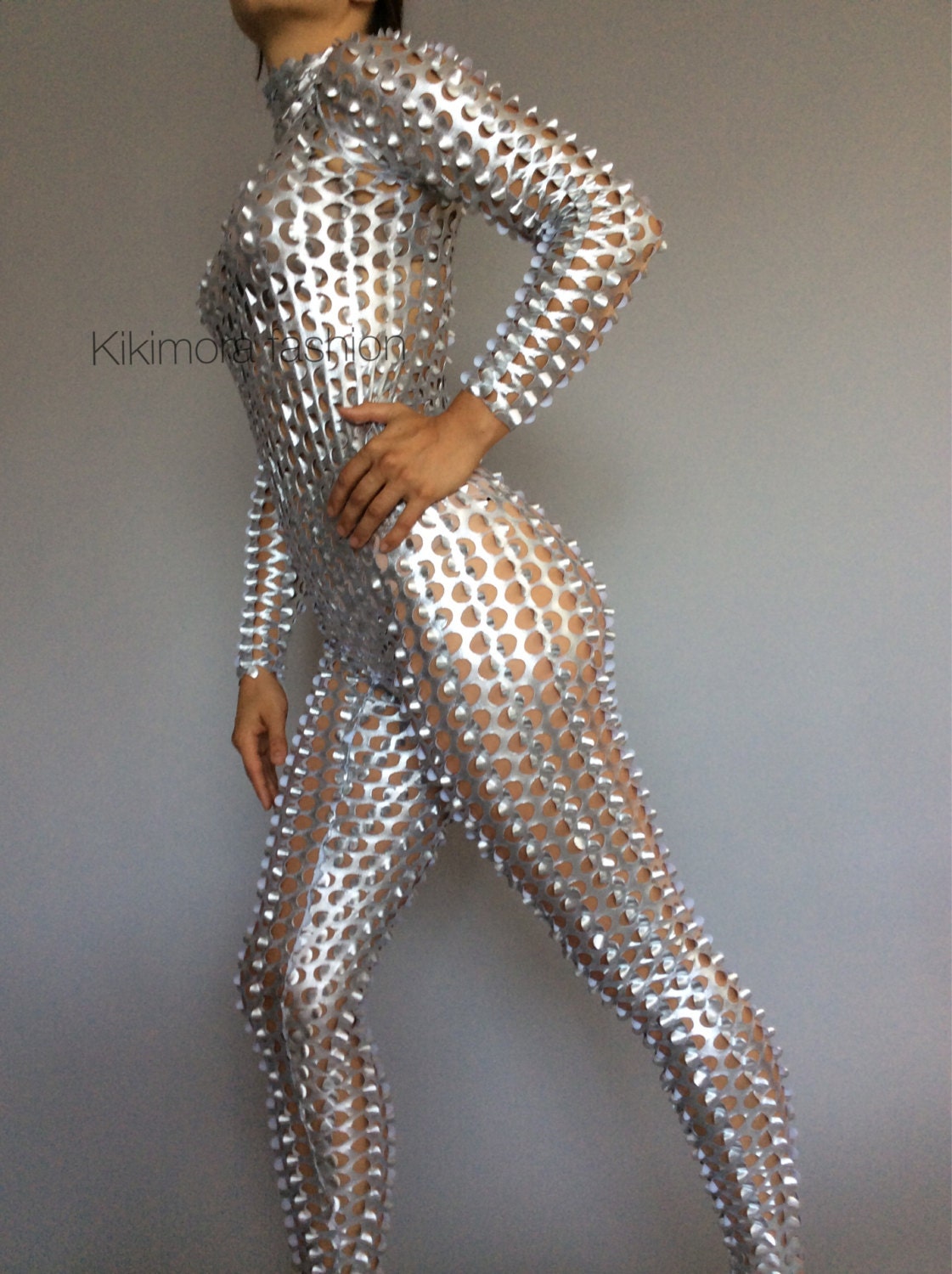 Person wearing a shiny, silver, full-body suit with circular cutout patterns, standing against a plain background.