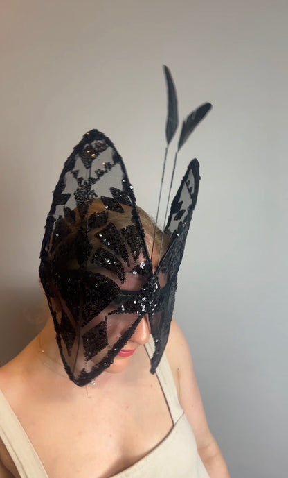 A person wears the Butterfly Headpiece—a festive black lace hat and open face headwear—making a striking holiday costume accessory, shown against a plain gray background.