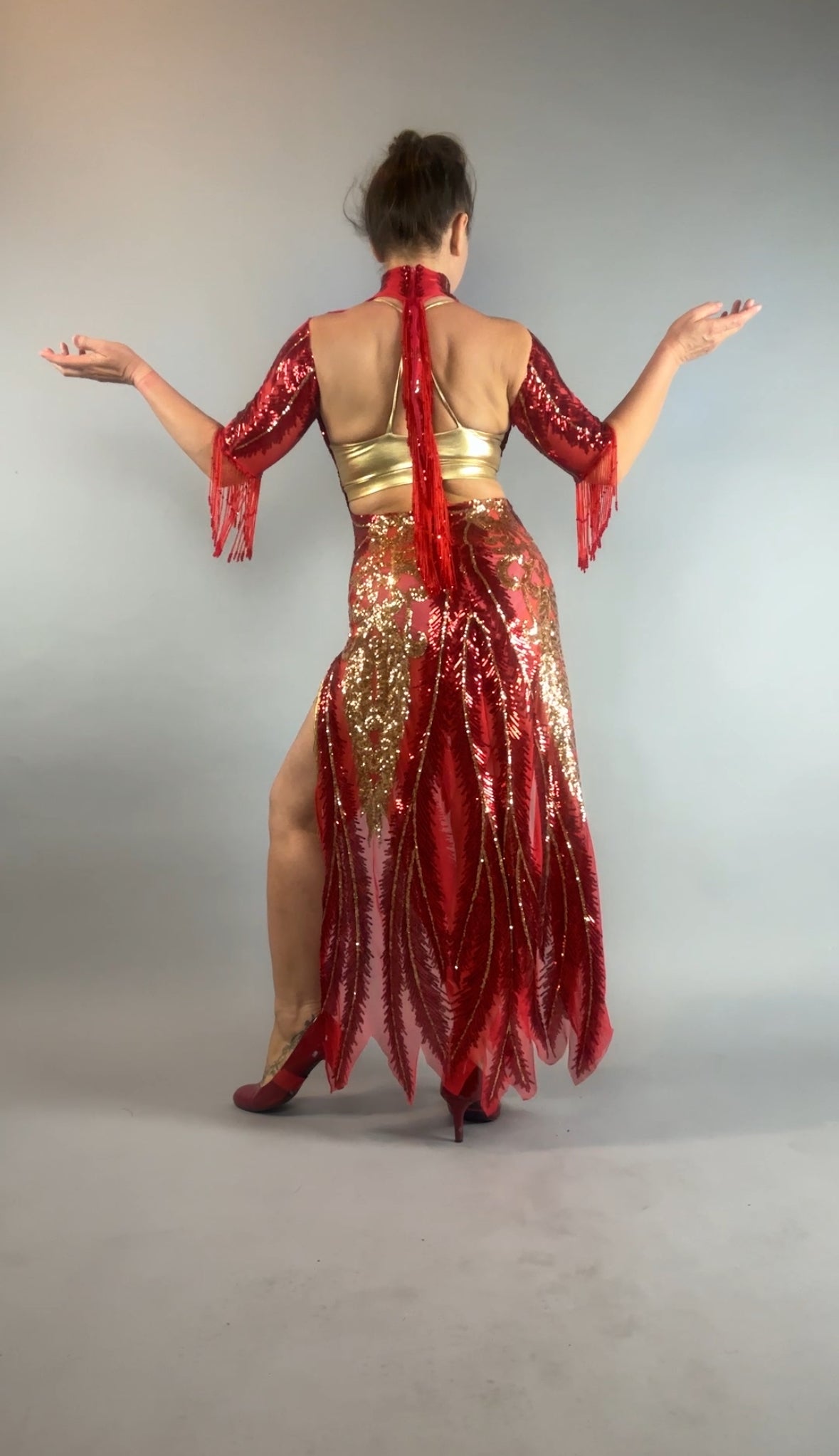 Model wearing the bearded French accessories Sequin Sheer Dress, featuring a backless design and beaded fringe accessories, poses with arms out and back to camera against a gray background.