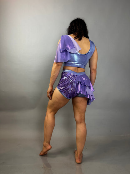 A person stands barefoot facing away, wearing a lavender purple Sequin Dance Costume featuring high waist shorts with half skirt, a crop top, and 4-way stretch mesh.