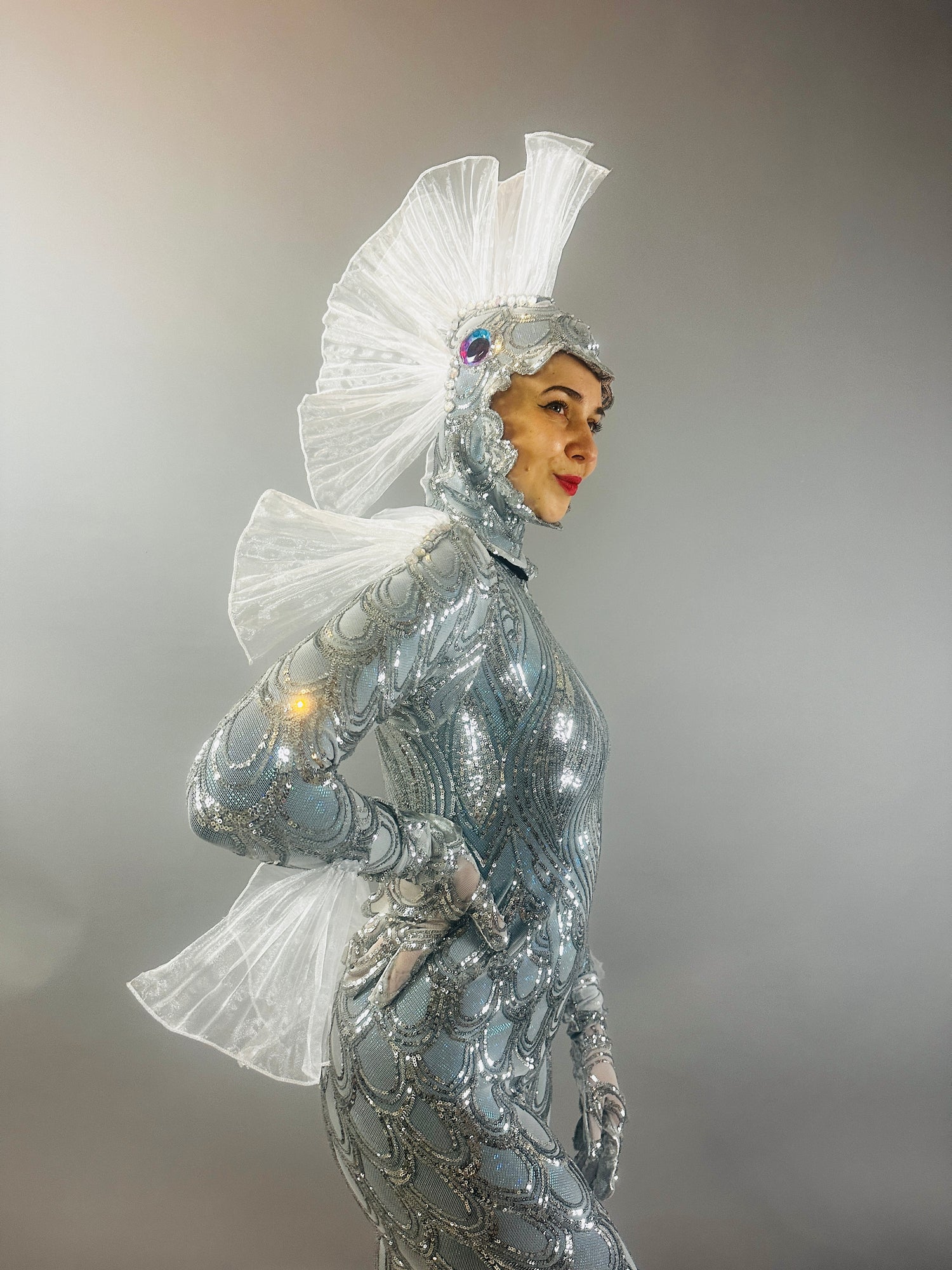 A person wears the Sea Creature—a shiny silver costume featuring fin-like headpiece and back fins—posing against a gray background, showcasing bold couture design.