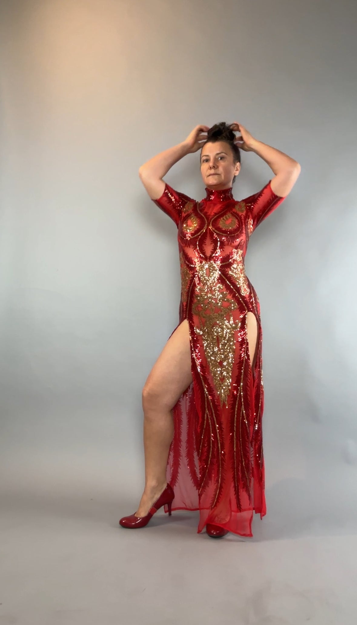 Woman wearing a Red And Gold Sequin Sheer Dress, sleeveless with hand accessories, poses confidently in heels against a plain gray background.