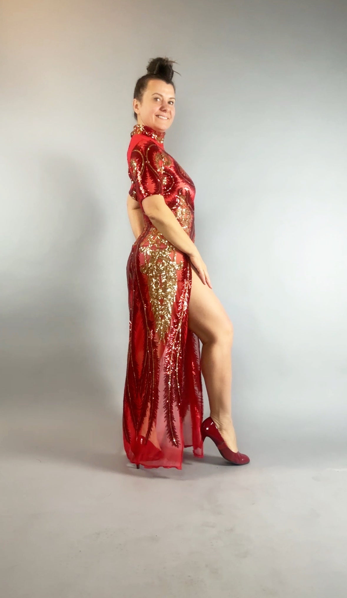 Woman wearing a Red and Gold Sequin Sheer Dress, sleeveless, accessorized with matching hand accessories, standing sideways and smiling.