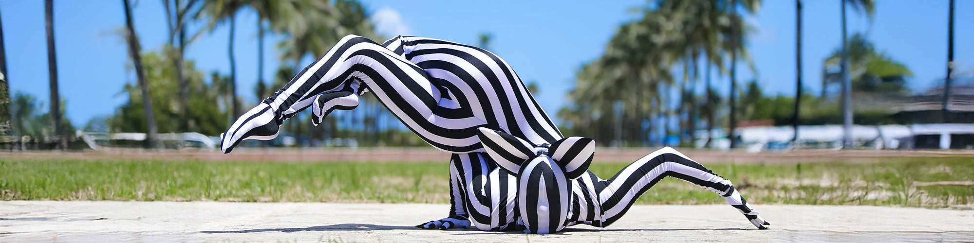 Two people in black and white striped bodysuits perform a contortion pose outdoors with palm trees behind them.