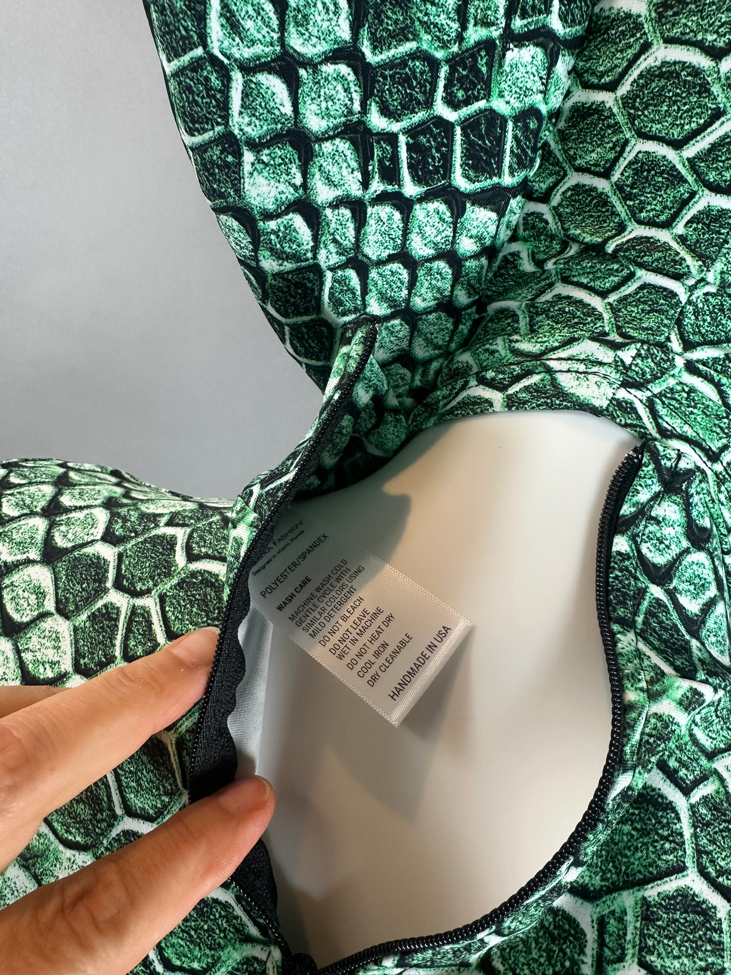 A hand opens the zipper of a green, scale-print fabric—part of the Green pixel Cobra Costume Snake Print Catsuit Serpent Cosplay—revealing a white tag with text inside.
