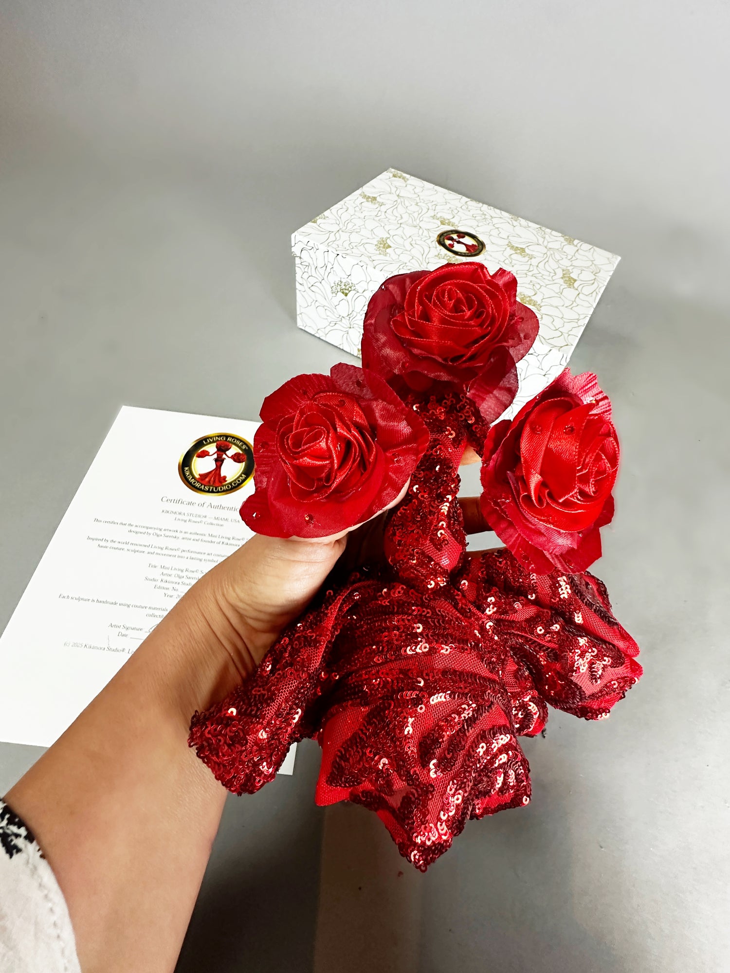 A hand holds the Mini Living Rose sculpture— a handcrafted, sequined doll ornament from Kikimora Studio—one of three magical art pieces, photographed with a white box and paper in the background.