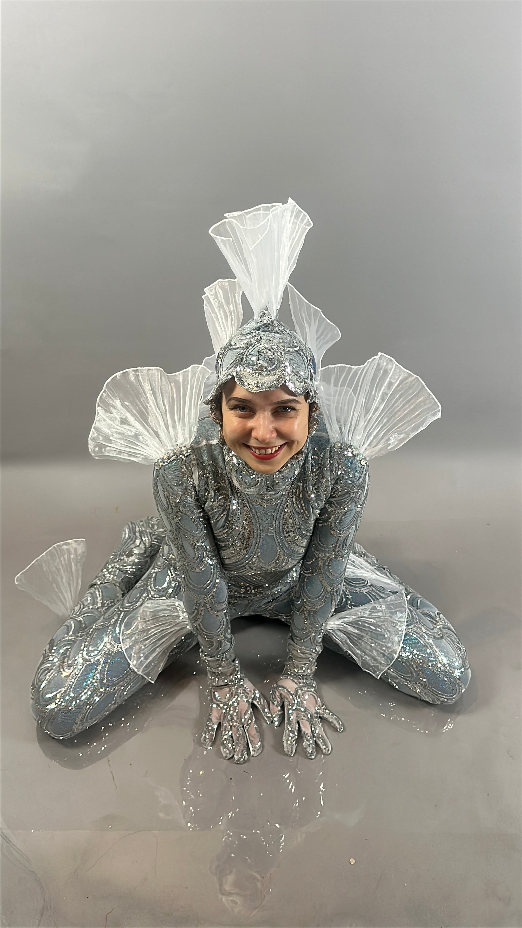 A person in the shiny, silver Sea Creature costume—with fins—crouches and smiles against a plain grey background, showcasing this playful wearable sculpture.