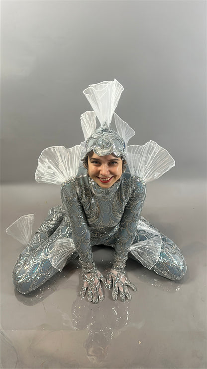 A person in the shiny, silver Sea Creature costume—with fins—crouches and smiles against a plain grey background, showcasing this playful wearable sculpture.