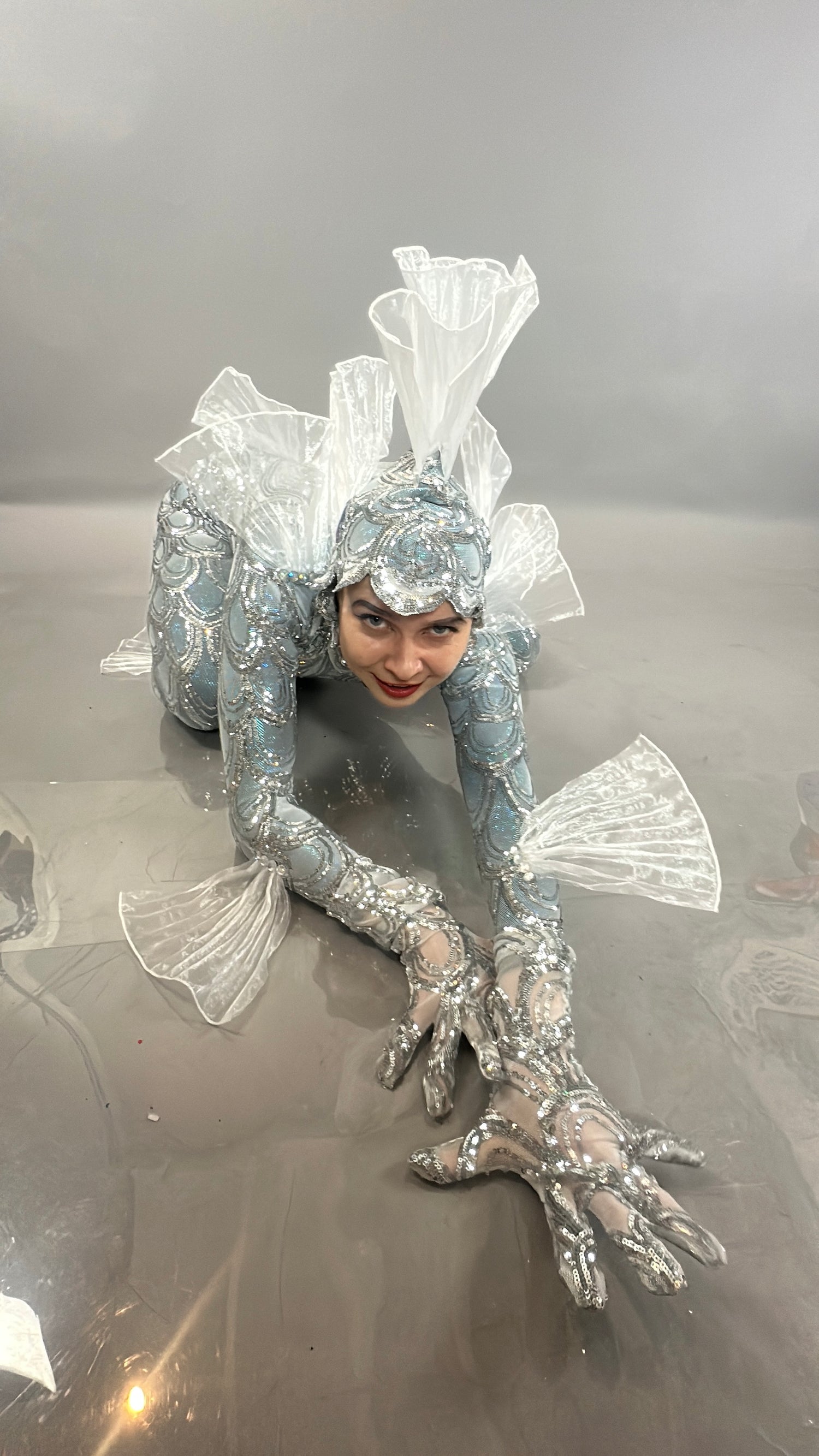 A person in the Sea Creature costume crouches on the floor, displaying translucent fins and silver scales in this striking wearable sculpture.
