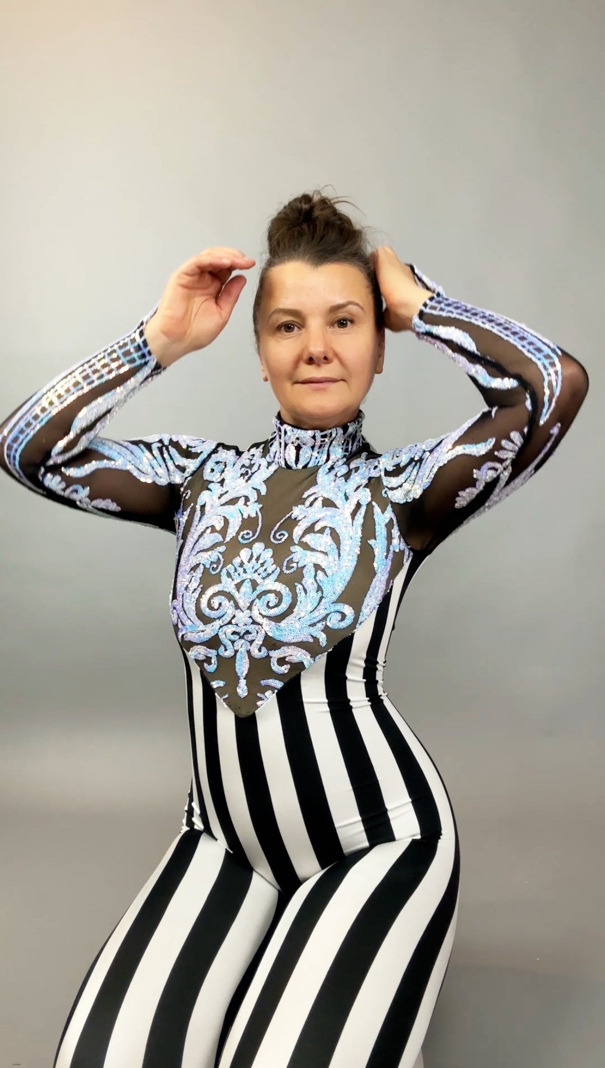 Woman in the Glamorous Sequins Stripes Catsuit costume, a black and white striped jumpsuit with blue ornate design, poses with hands near her head.