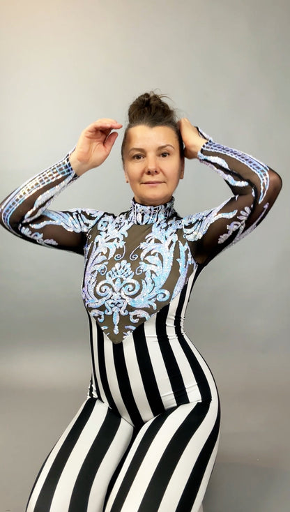 Woman in the Glamorous Sequins Stripes Catsuit costume, a black and white striped jumpsuit with blue ornate design, poses with hands near her head.