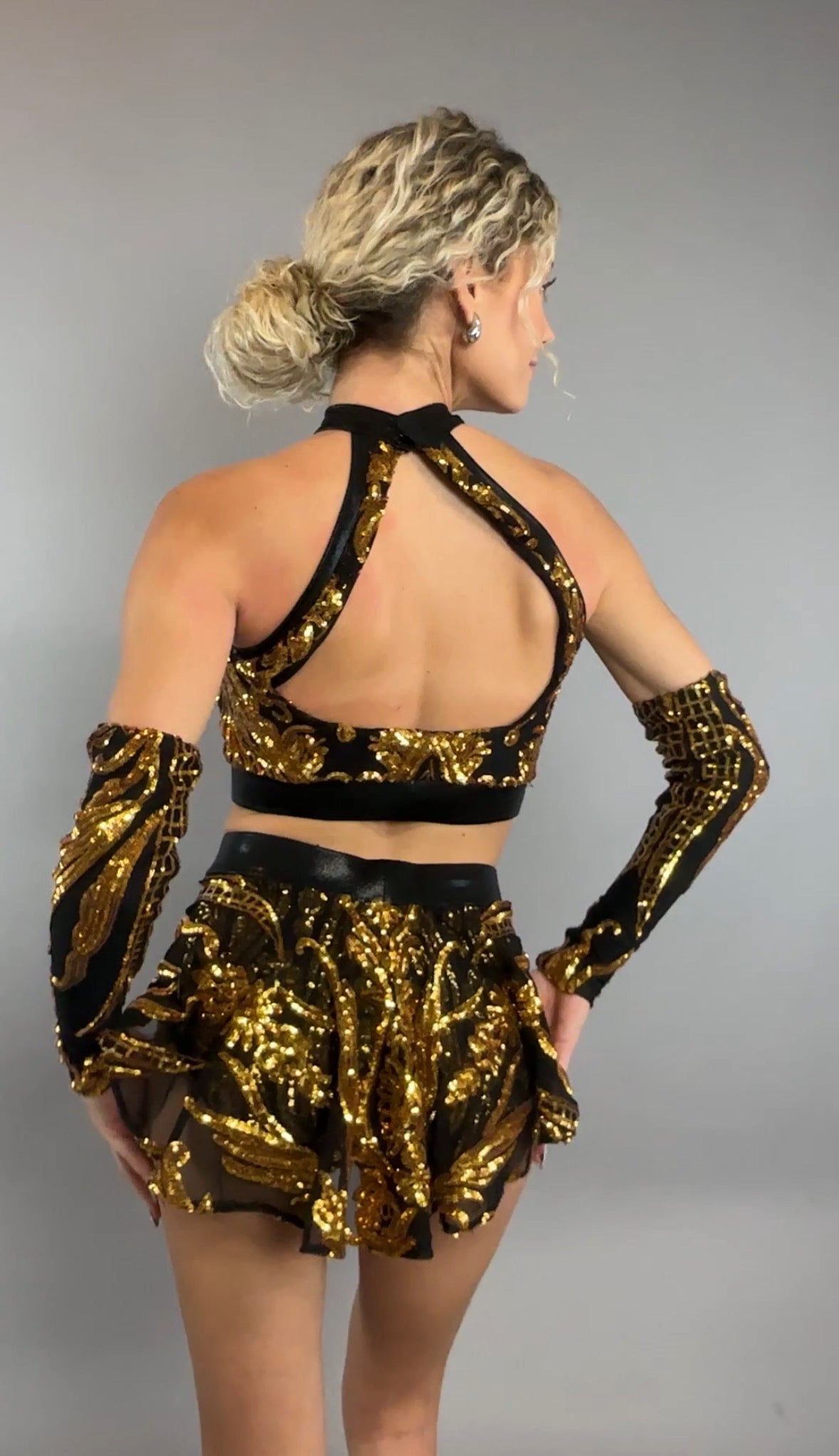 A woman models the Gold Sequin on Black Crop Top and Bottom Set, featuring a halterneck top and matching hand accessories with delicate black mesh accents, shown from the back.