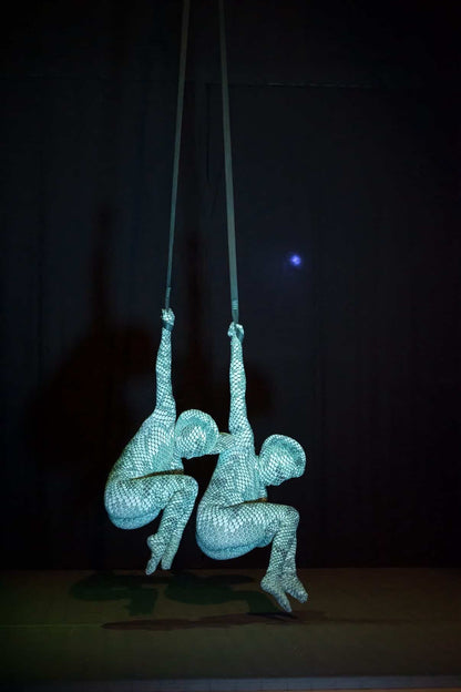 Two aerial performers wear custom-made Green Pixel Cobra snake print catsuits and headdresses, hanging upside down on straps against a dark background for a striking contortionist or serpent cosplay display.