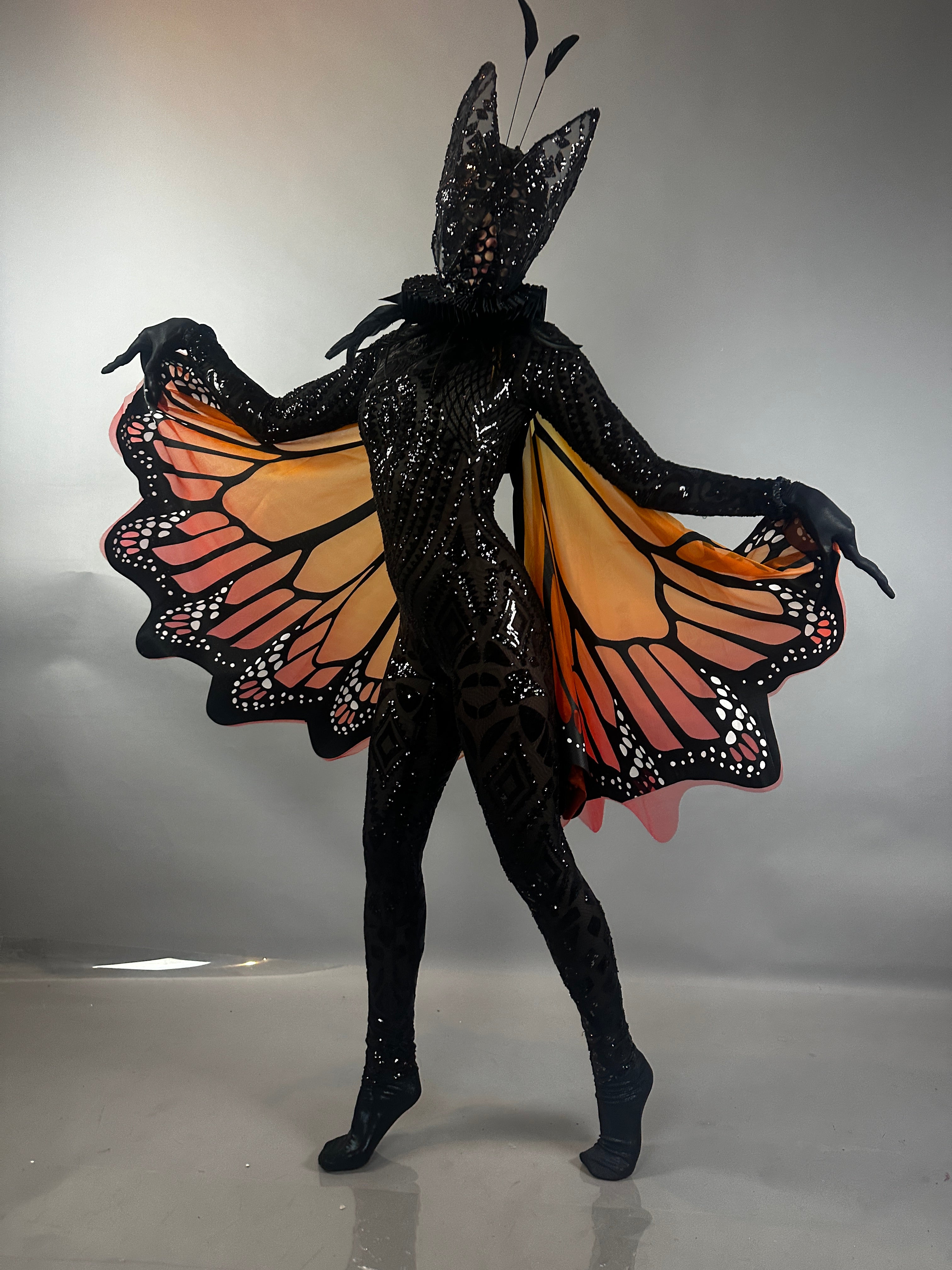 A dramatic pose features the Black Sequin Full Costume Butterfly—an elegant black bodysuit with vibrant orange wings and a matching headpiece—showcasing stunning wearable art.