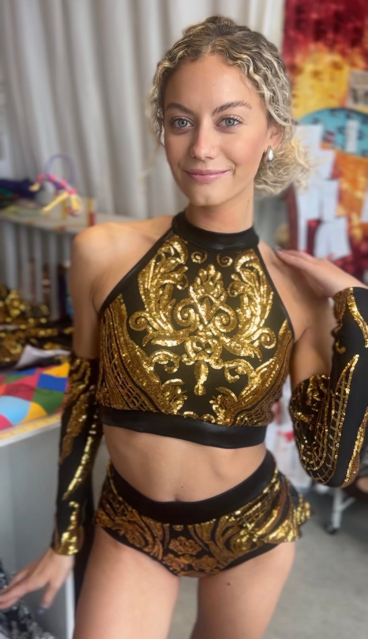 A woman poses indoors, smiling at the camera, wearing a Gold Sequin on Black Crop Top and Bottom Set featuring a halterneck top and matching hand accessories.