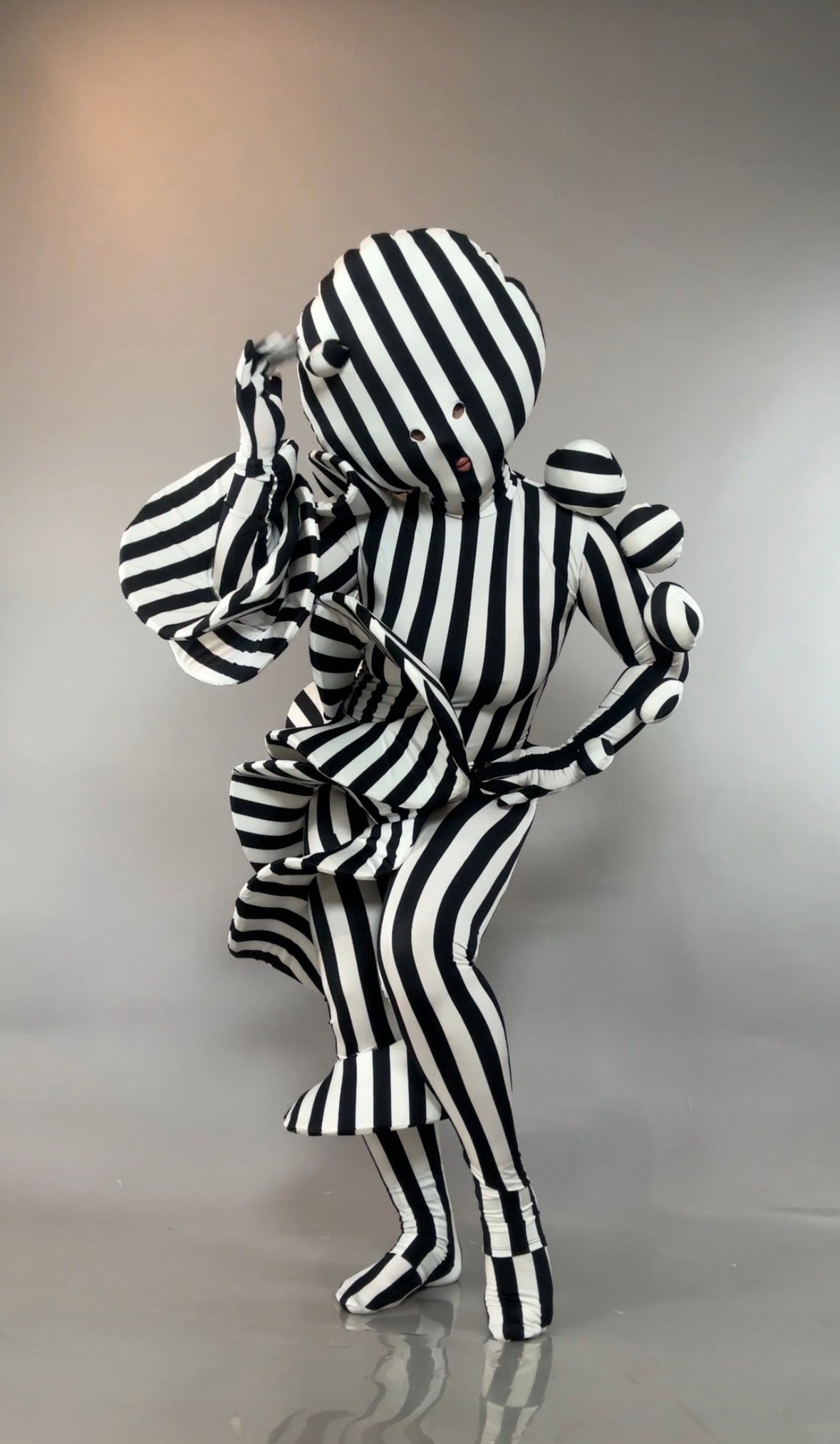 A person wears a Black and White Striped Creature Costume with sculptural spheres, a mask, gloves, and a ruffled hem.