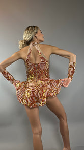 A person models the Fire color sparkling Halter Leotard, featuring red and gold sequin accents and detachable sleeves, with an asymmetrical skirt, posing with bent arms and facing away from the camera.