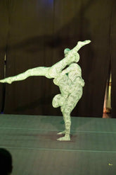 Two performers in matching Green Pixel Cobra Costumes—snake print catsuits with headdresses—present a balancing act on stage against a dark backdrop.