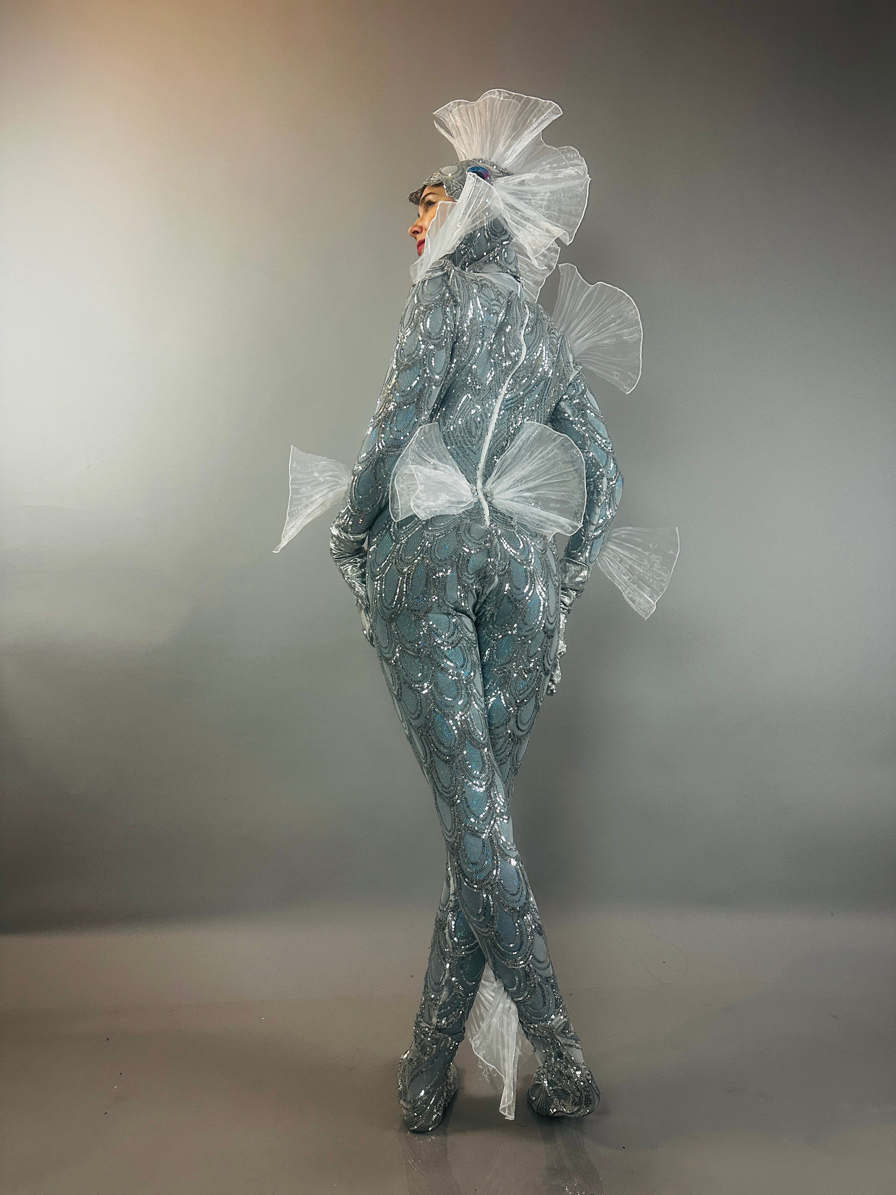 Model wears the Sea Creature costume—a shiny silver fish-scale bodysuit with translucent fin-like accents—posing against a gray background and highlighting avant-garde, couture-inspired design.