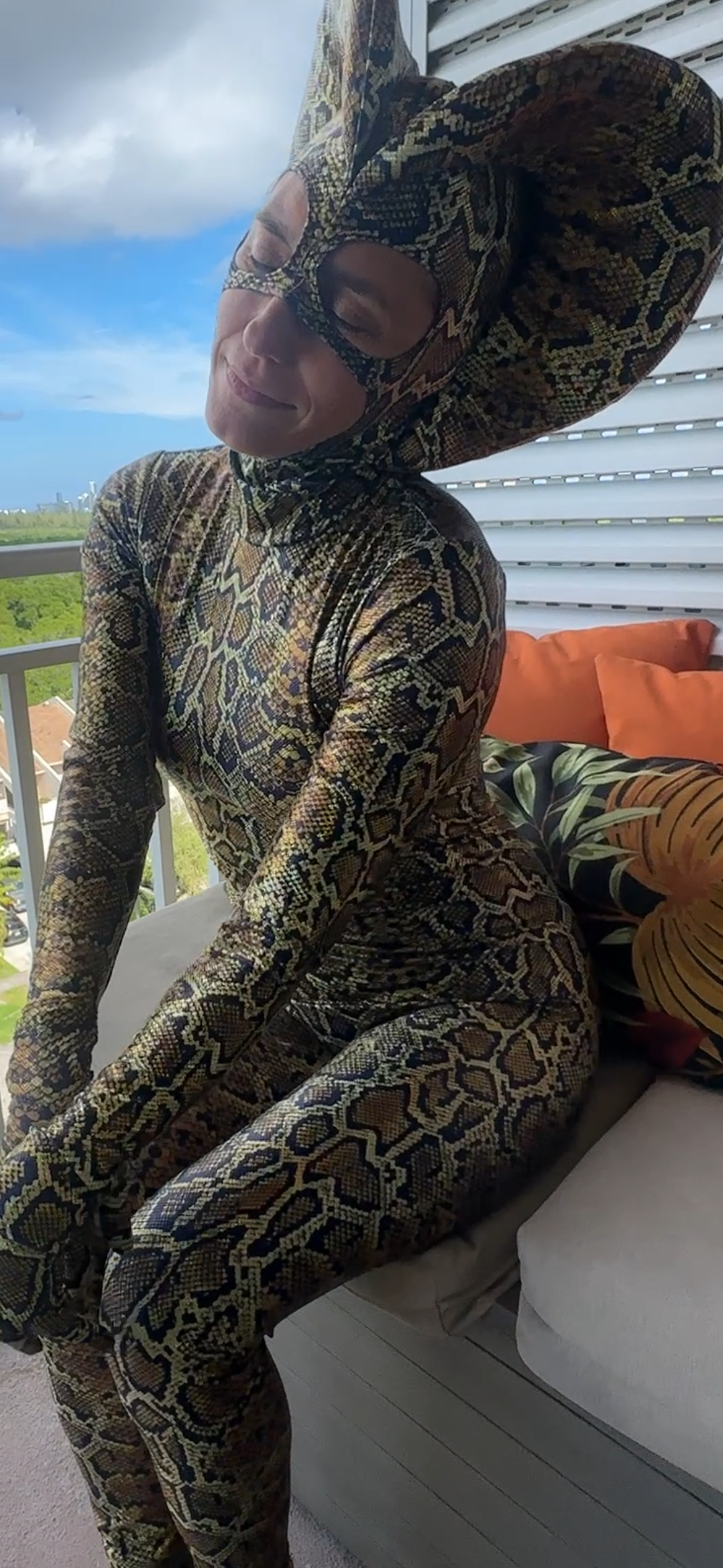 A person in the Green Cobra Snake Costume—custom-made for gymnastics, acrobatics, dancers, and contortionists—wears a striking snake headdress while sitting on a balcony couch decorated with orange pillows.