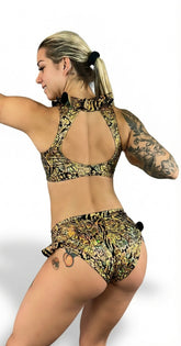 Woman with blonde hair poses with arms raised, showing back tattoos in the Gold & Black Snakeskin Crop Top & Bottom Set featuring an open back and cheeky bottoms.