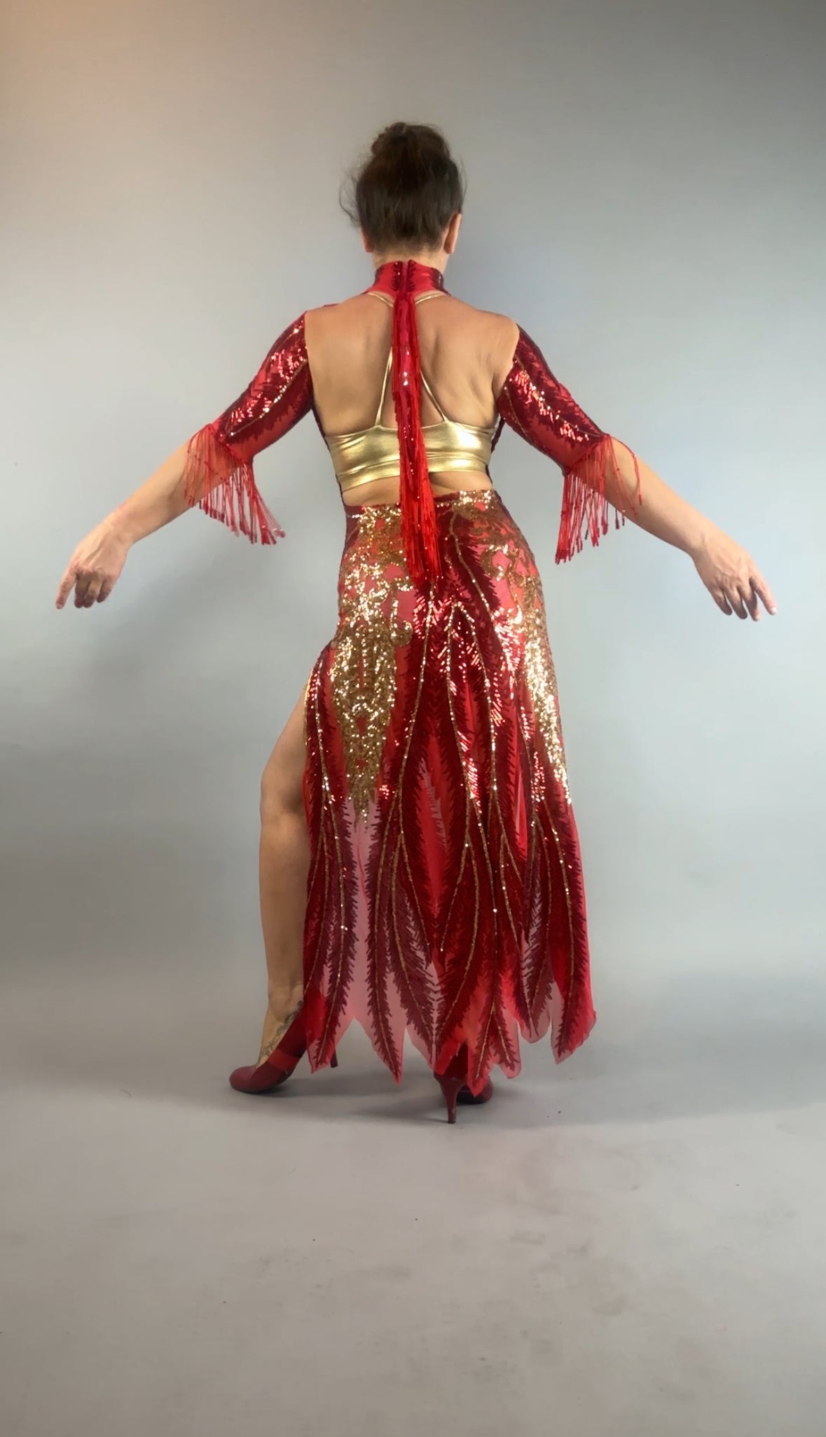 A woman in the bearded French accessories Sequin Sheer Dress—backless with beaded fringe—stands with arms outstretched, facing away.