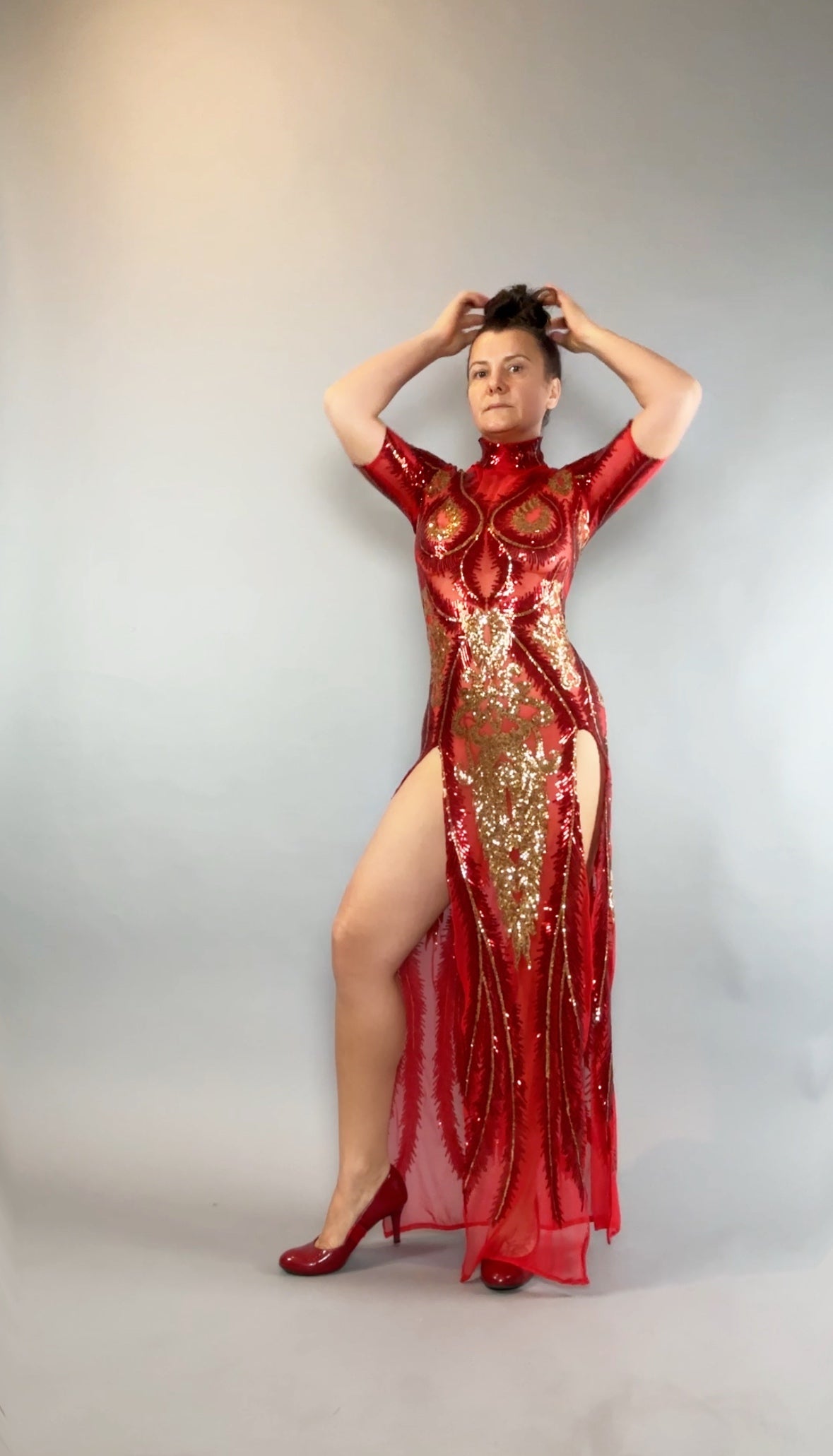 Model wears the Red And Gold Sequin Sheer Dress, sleeveless with a high side slit and hand accessories, posing against a plain gray background.