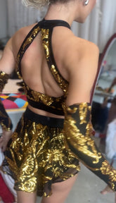 A person wearing the Gold Sequin on Black Crop Top and Bottom Set, featuring a halterneck top, hand accessories, golden sequins, and an open-back design, stands near a mirror and colorful fabric.
