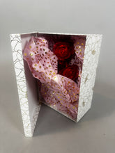 A white floral box partially open reveals the Mini Living Rose sculpture doll ornament by Kikimora Studio—an exquisite handcrafted red rose wrapped in pink tissue paper with gold dots inside.