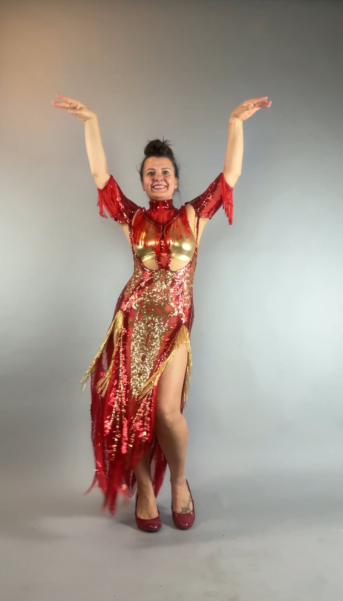 Woman wears the bearded French accessories Sequin Sheer Dress—backless, with beaded fringe—posing with arms raised and smiling against a plain gray background.