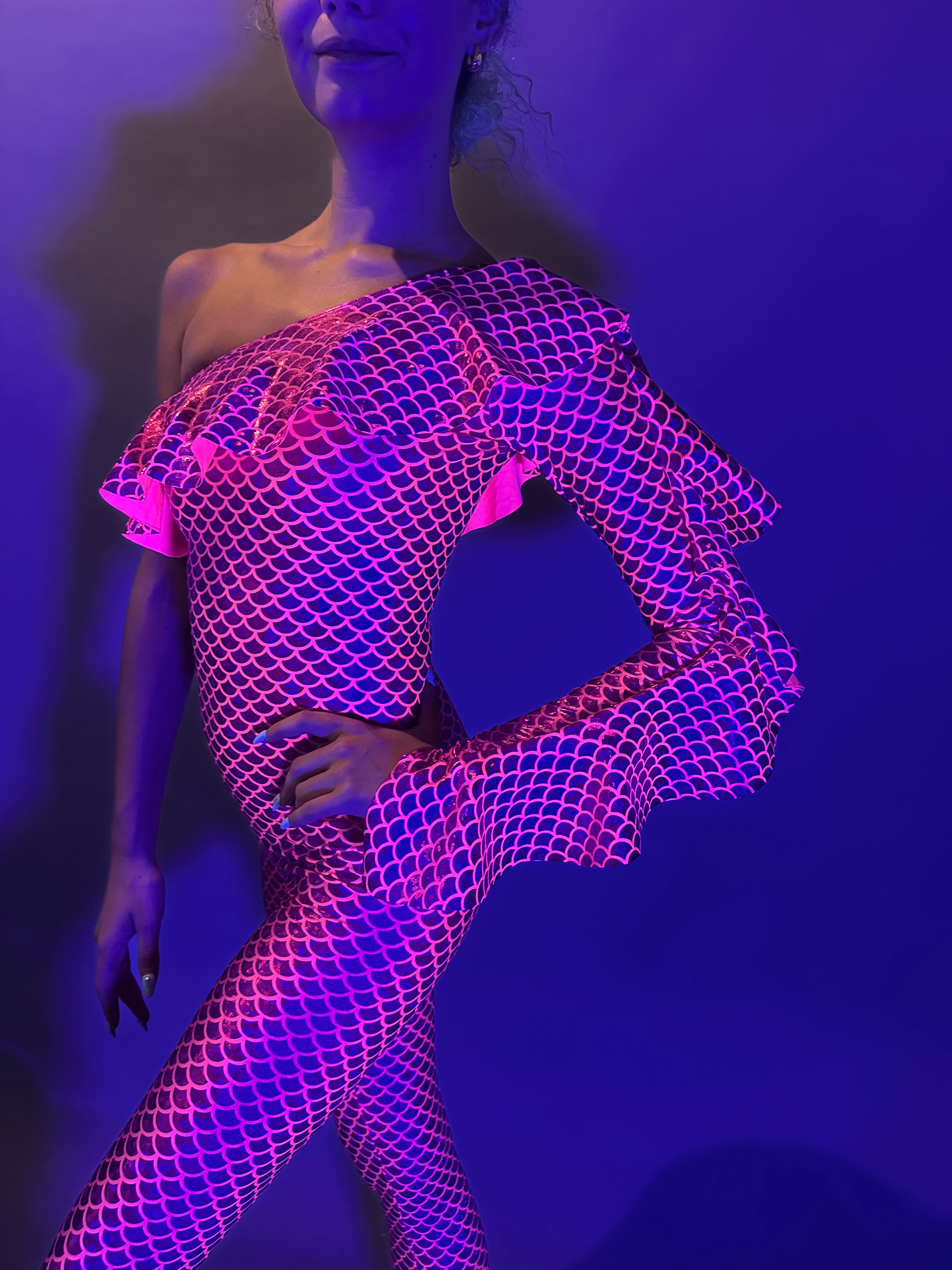 Person poses under dramatic purple lighting wearing a Red Ruffle Catsuit—an off-shoulder, asymmetrical spandex bodysuit with ruffle design, available in custom colors.