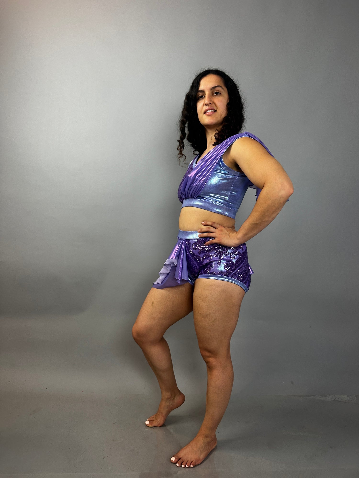 A person poses barefoot in a lavender purple Sequin Dance Costume featuring high waist shorts with a half skirt, crop top, and 4-way stretch mesh against a plain gray background.