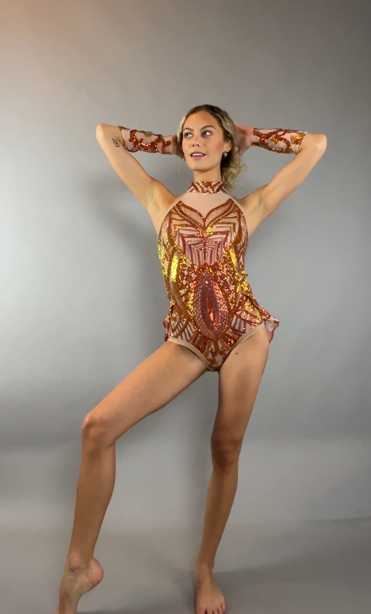 A woman poses against a plain gray background, looking to the side, wearing the Fire color sparkling Halter Leotard with sequin accents and detachable sleeves.