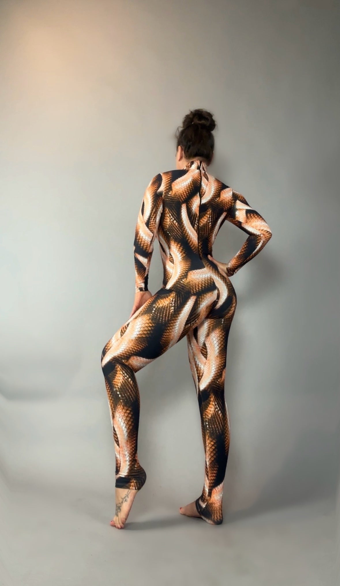 Person in a Brown Snake print Catsuit with turtleneck and long sleeves poses barefoot against a plain, light gray background, facing away.