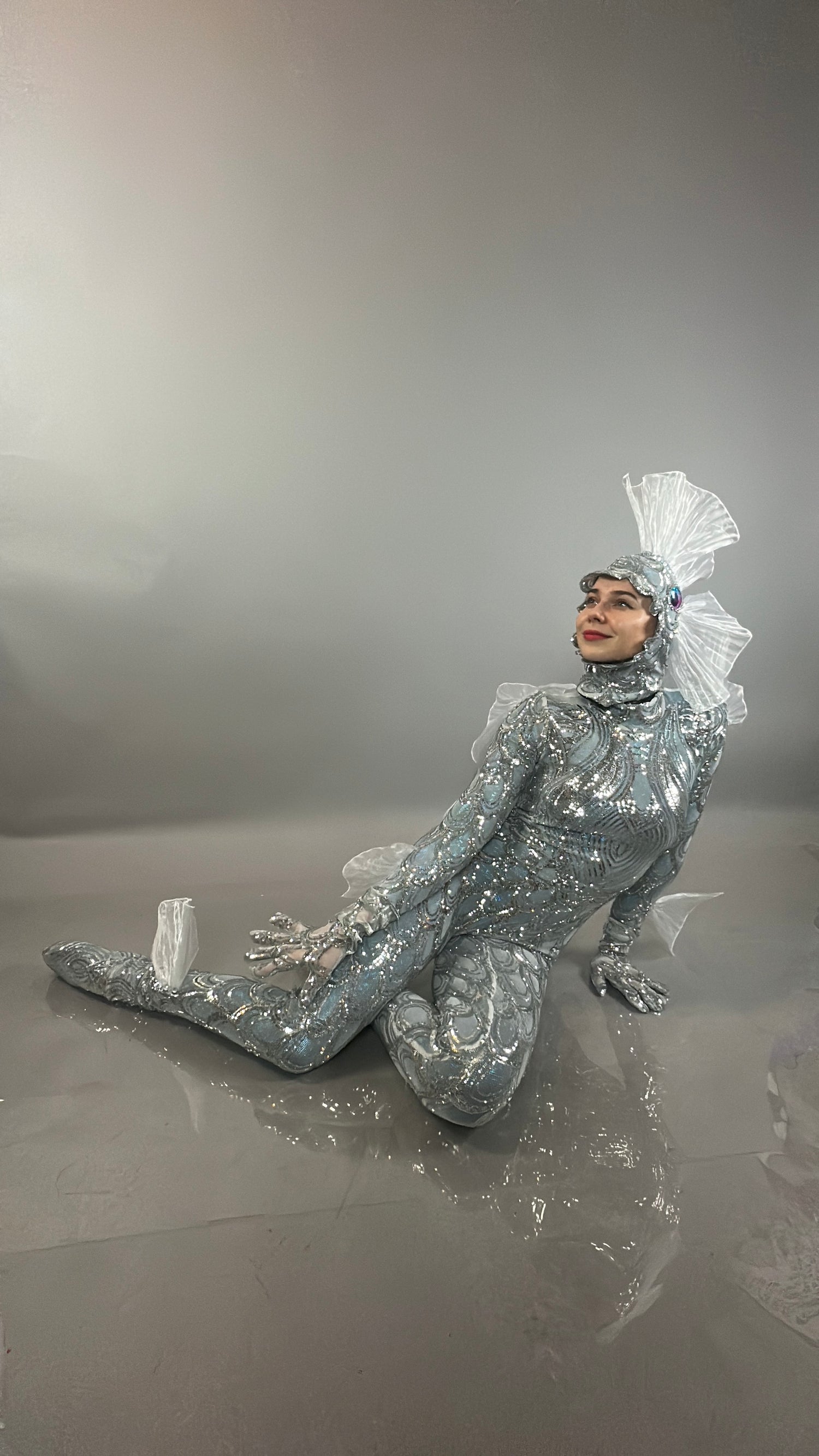 Model in the Sea Creature bodysuit with an elaborate headpiece poses seated on the floor against a gray backdrop, capturing the striking allure of couture-inspired design.