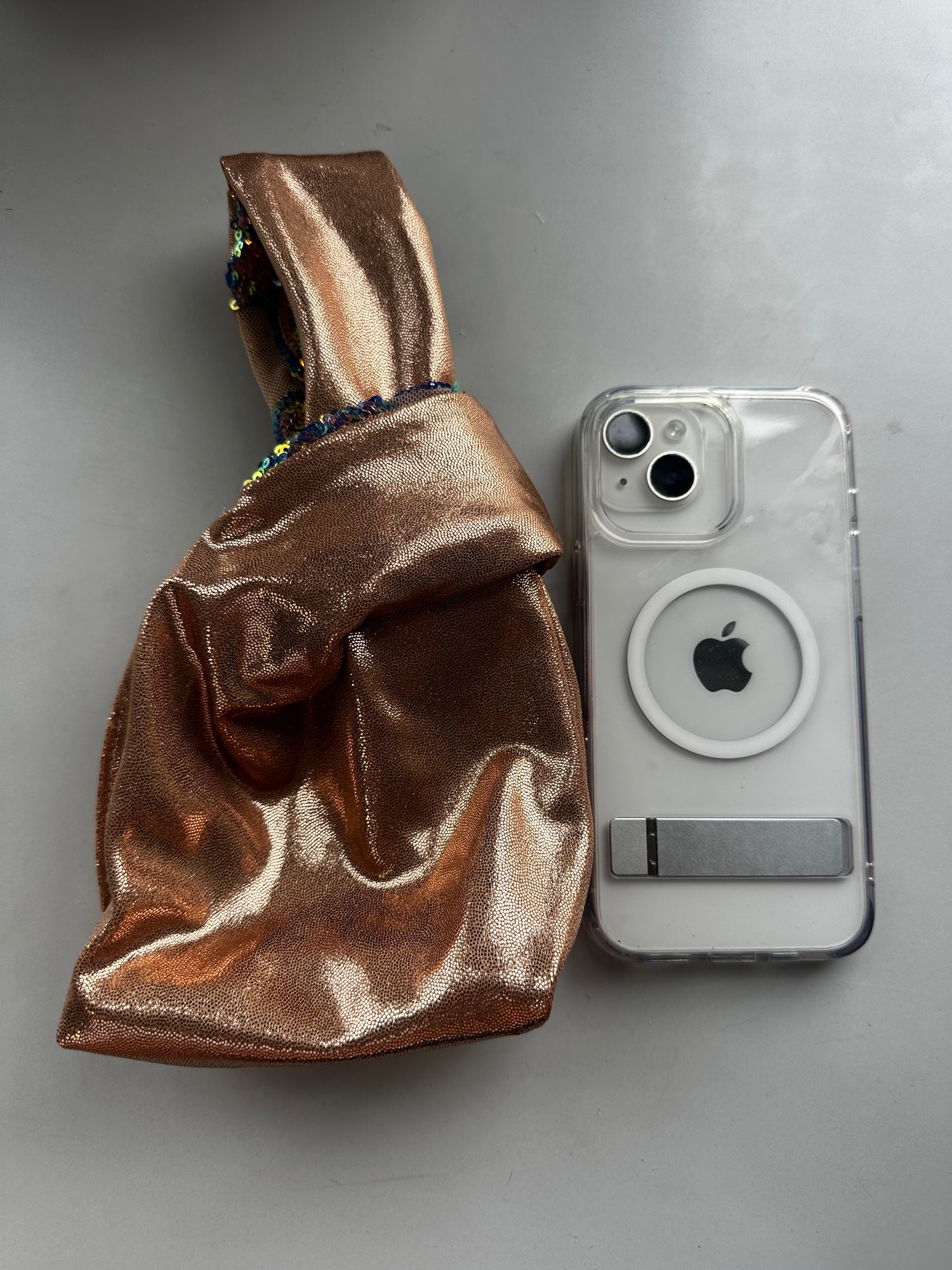 A handcrafted reversible sequin mini tote—an eco-friendly fashion accessory—rests beside a white iPhone with a clear MagSafe case on a gray surface.