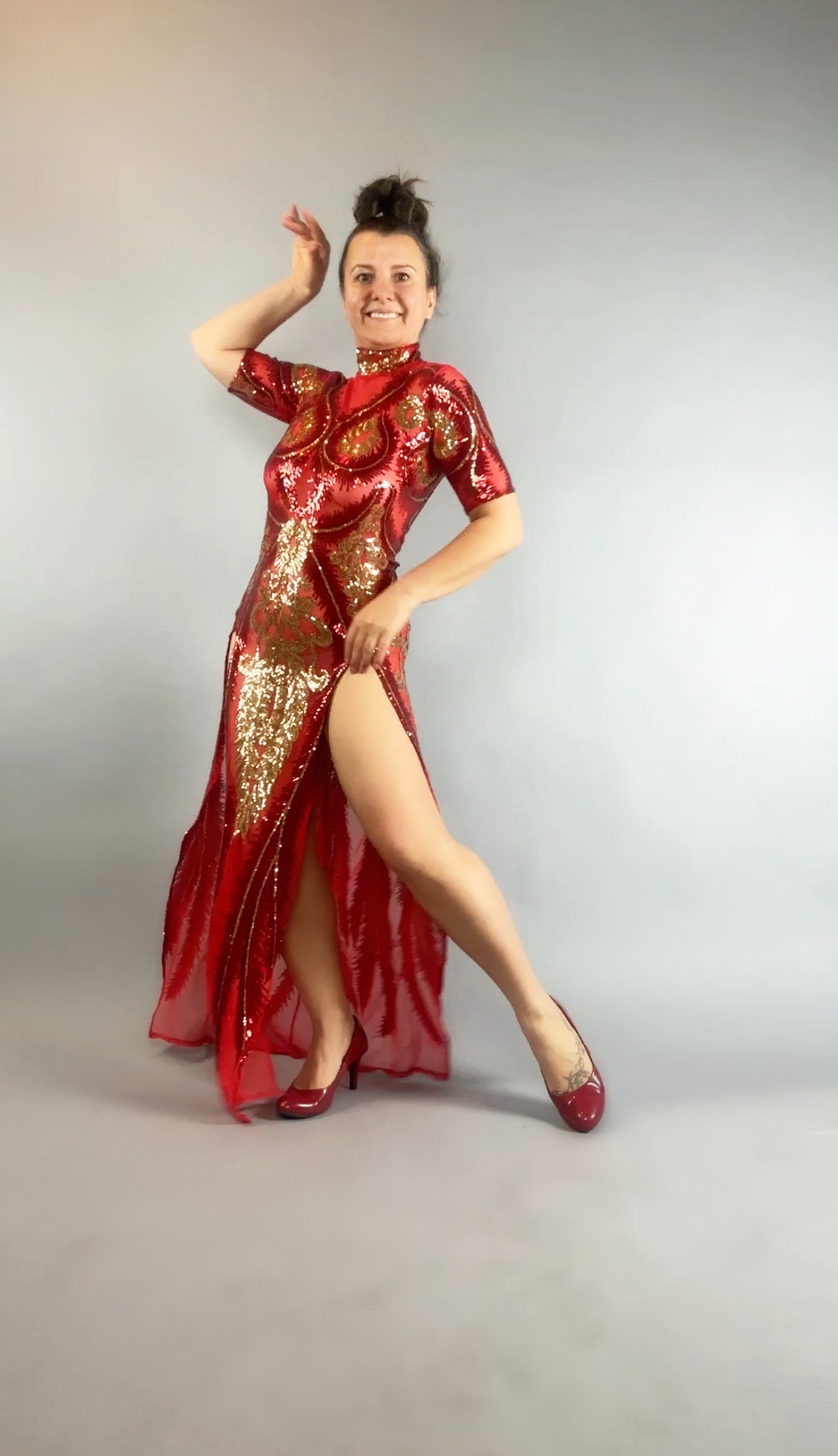 Woman wears a Red and Gold Sequin Sheer Dress, sleeveless, with hand accessories, posing with one leg raised and smiling against a plain light background.
