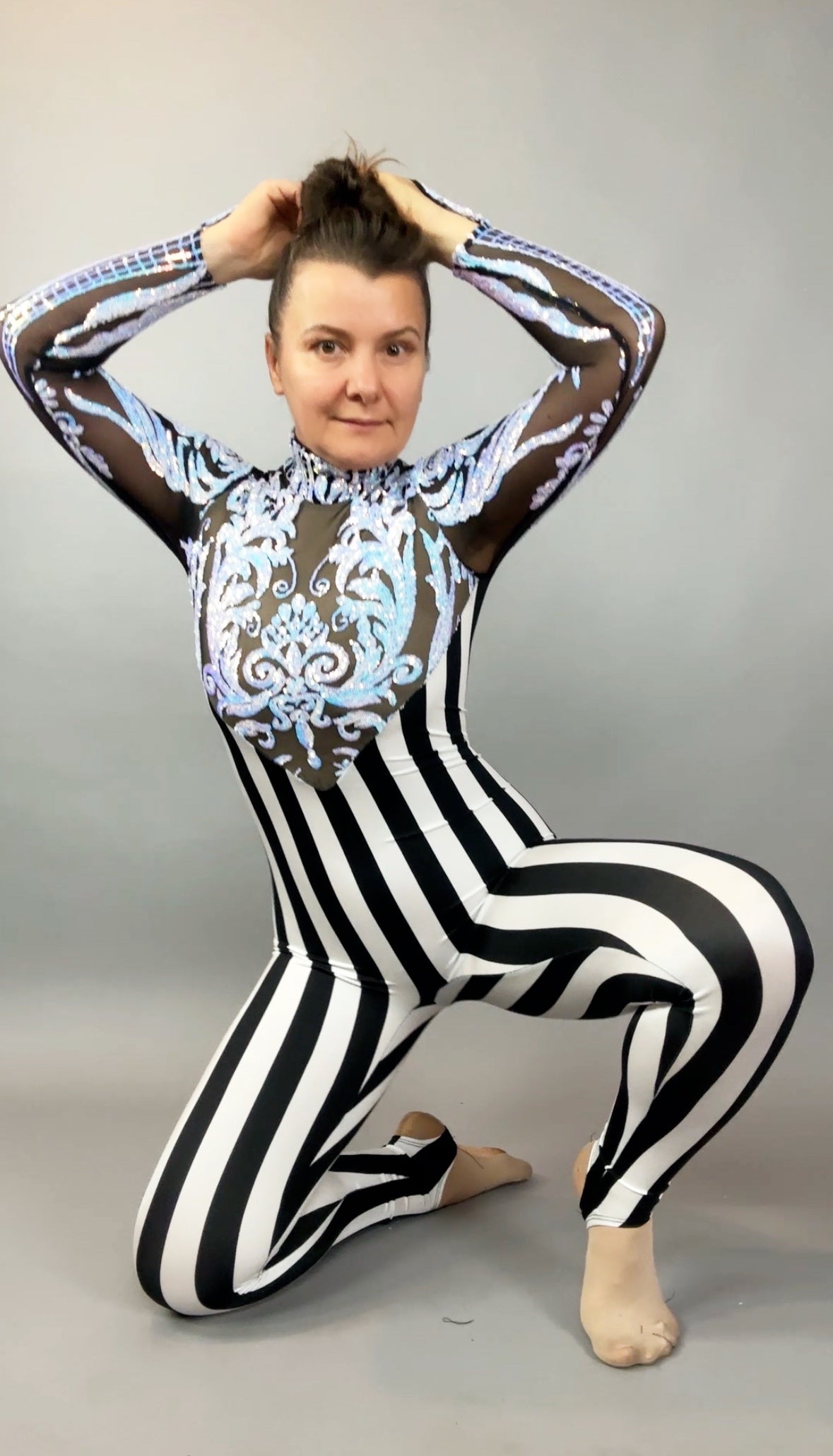 Person kneeling in the Glamorous sequins Stripes Catsuit costume, featuring black-and-white stripes, blue patterns, and a sequined leaf design, hands holding hair, on a gray background.