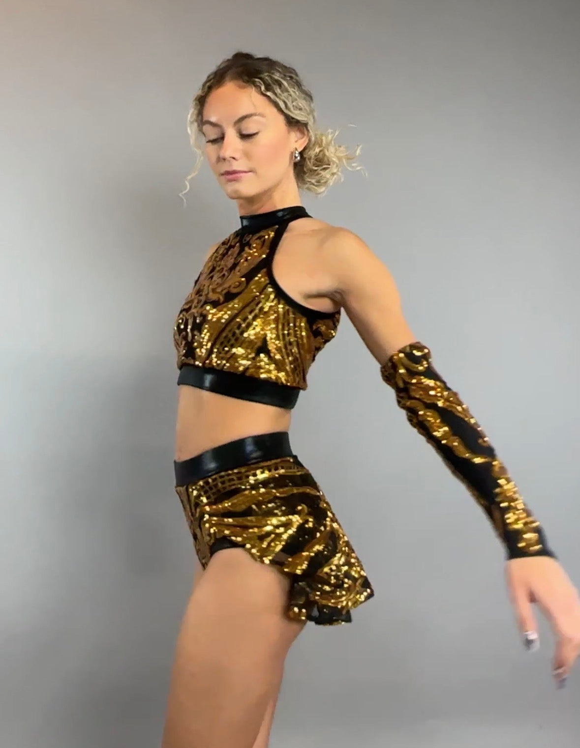 A woman models the Gold Sequin on Black Crop Top and Bottom Set, featuring a halterneck top and hand accessories, against a plain gray background.