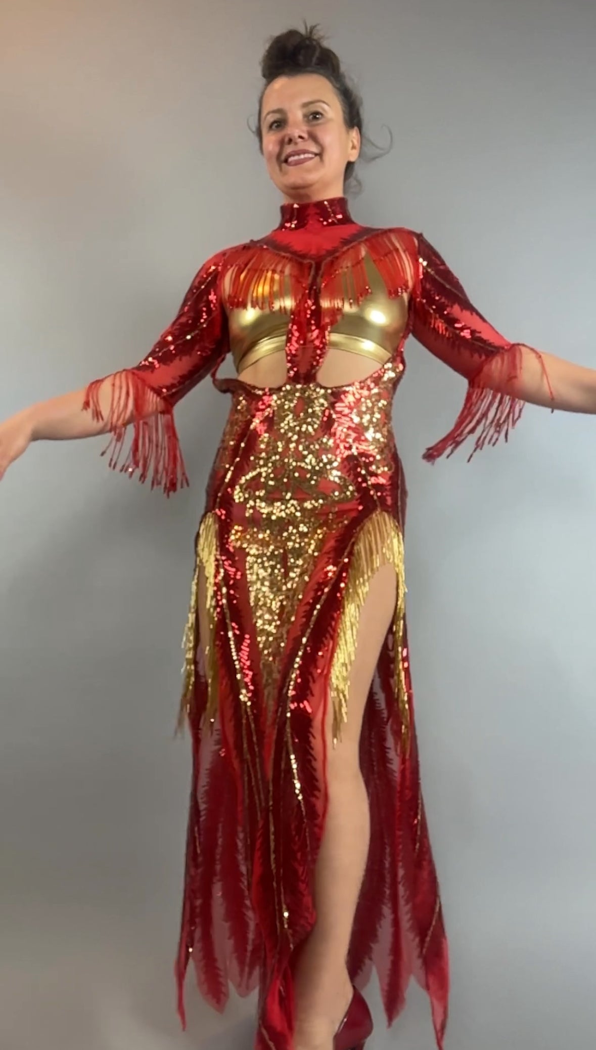 A woman poses with arms outstretched in the bearded French accessories Sequin Sheer Dress—backless and adorned with beaded fringe accessories—against a plain gray background, radiating glamour.