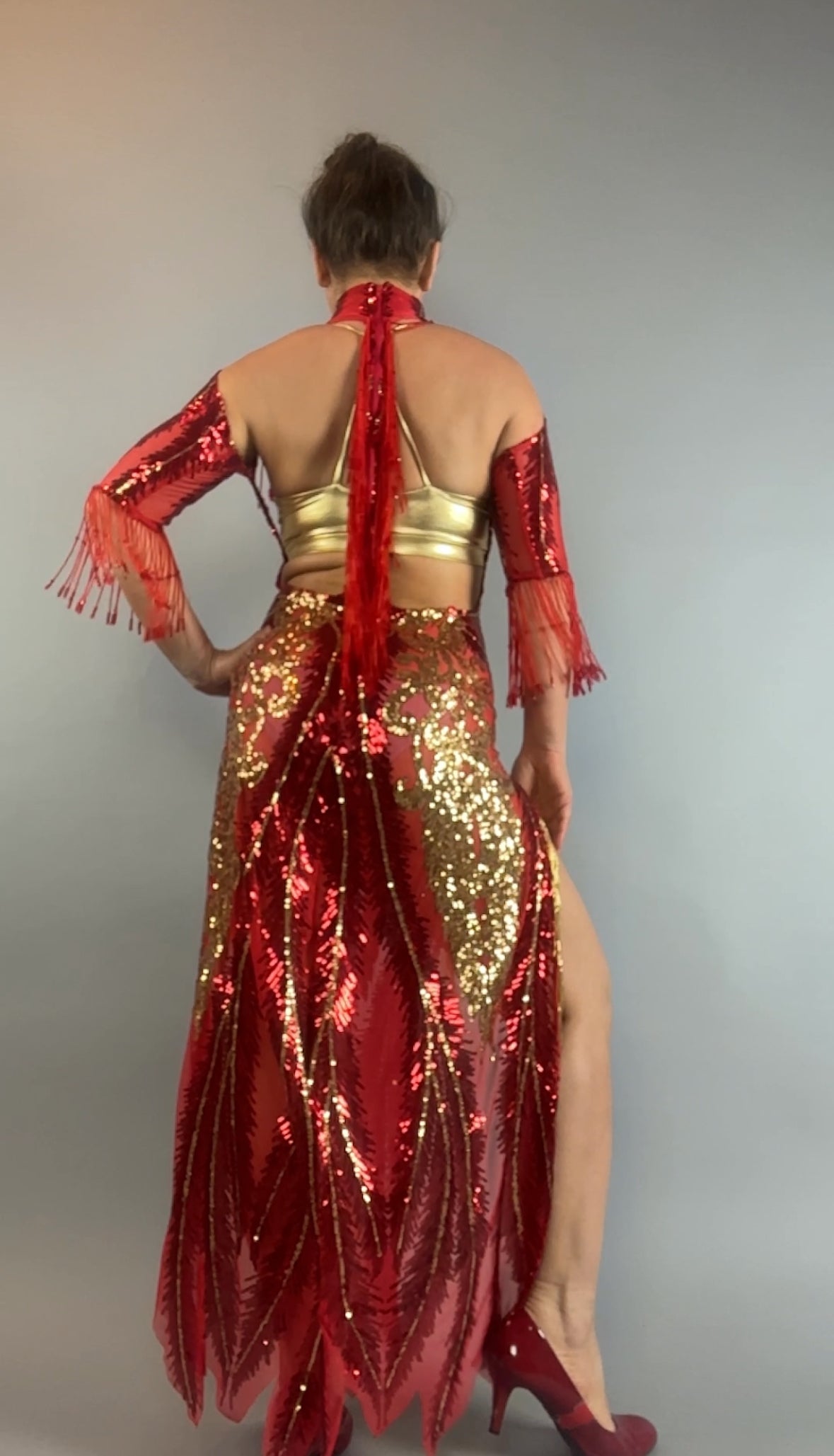 Person wearing the bearded French accessories Sequin Sheer Dress, a backless red maxi with gold beaded fringe, standing with their back to the camera against a gray background.