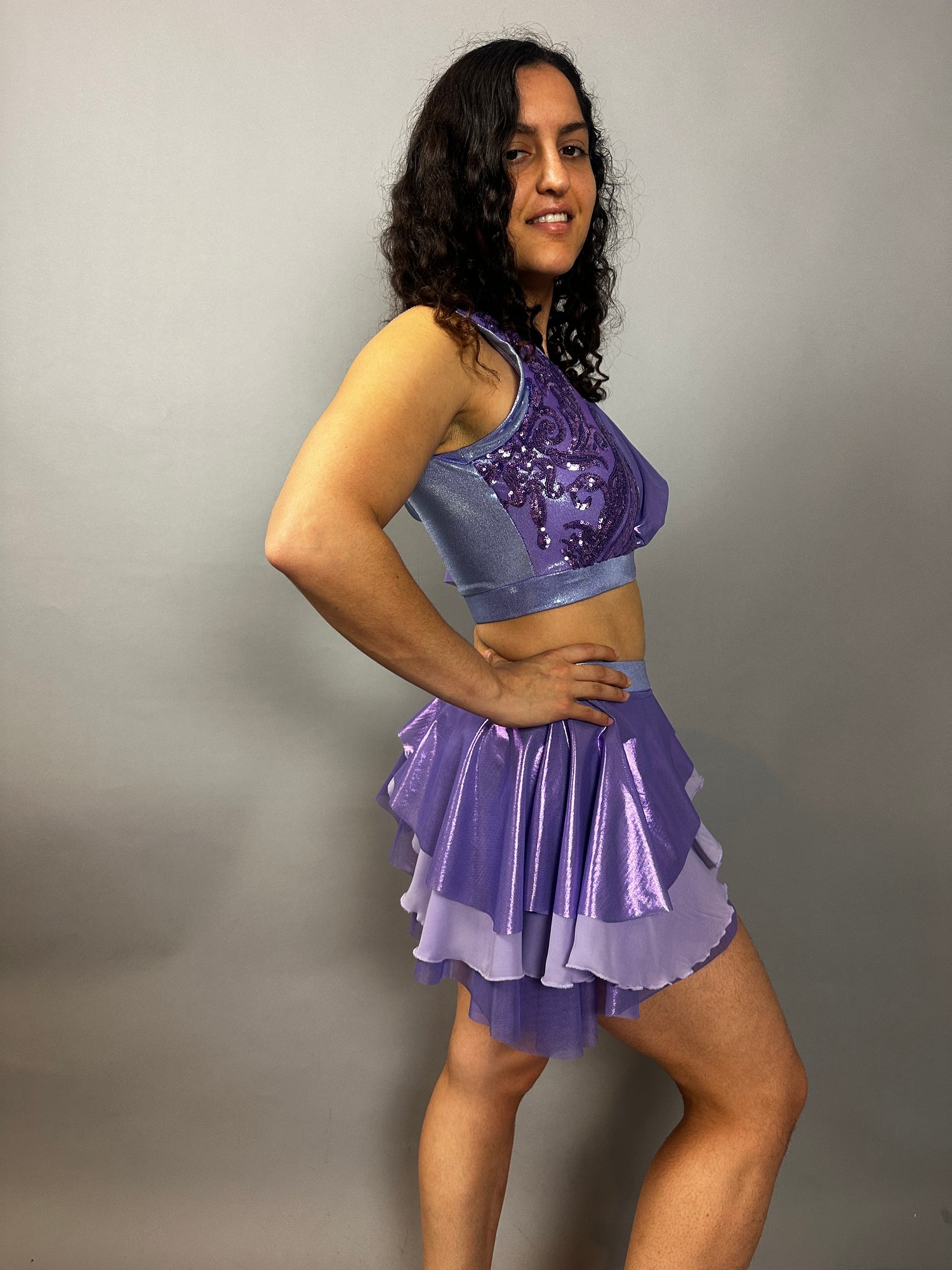 A woman poses sideways against a plain gray background, modeling the Lavender Purple Sequin Dance Costume featuring a crop top, high waist shorts with a half skirt, and 4-way stretch mesh—an eye-catching fire dancer outfit.