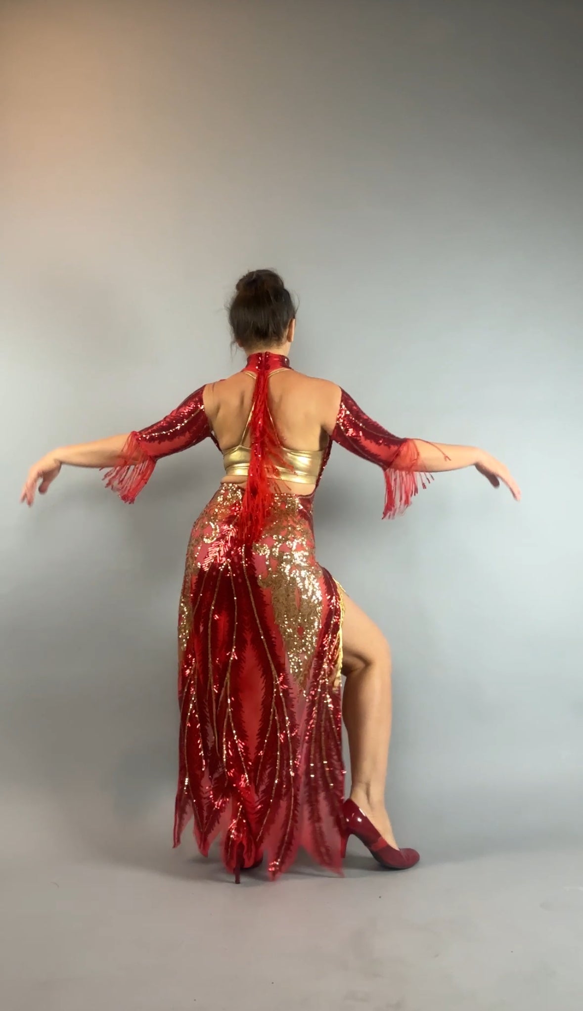 A woman poses with arms out, seen from the back against a gray background, wearing the bearded French accessories Sequin Sheer Dress—backless with beaded fringe accessories.