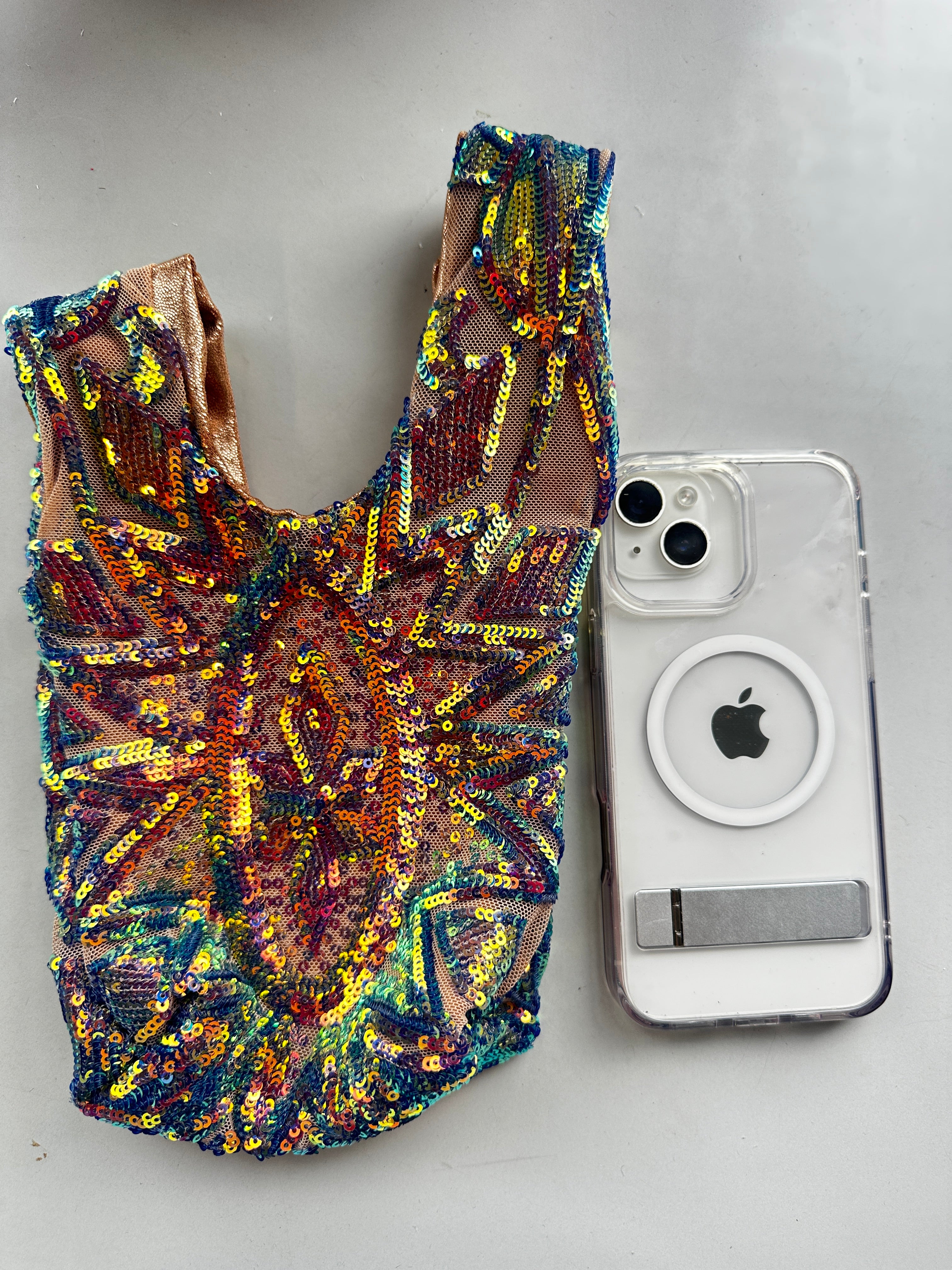 A colorful, sequined doll dress sits beside a clear iPhone case with a MagSafe ring on a gray surface, paired with a handcrafted reversible sequin mini tote—a sustainable fashion accessory.
