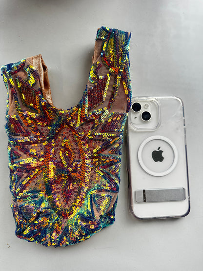 A colorful, sequined doll dress sits beside a clear iPhone case with a MagSafe ring on a gray surface, paired with a handcrafted reversible sequin mini tote—a sustainable fashion accessory.
