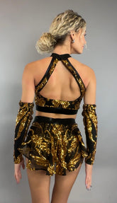 A woman is seen from the back against a gray background, wearing the Gold Sequin on Black Crop Top and Bottom Set featuring a halterneck top with golden sequins, black mesh accents, and coordinating hand accessories.
