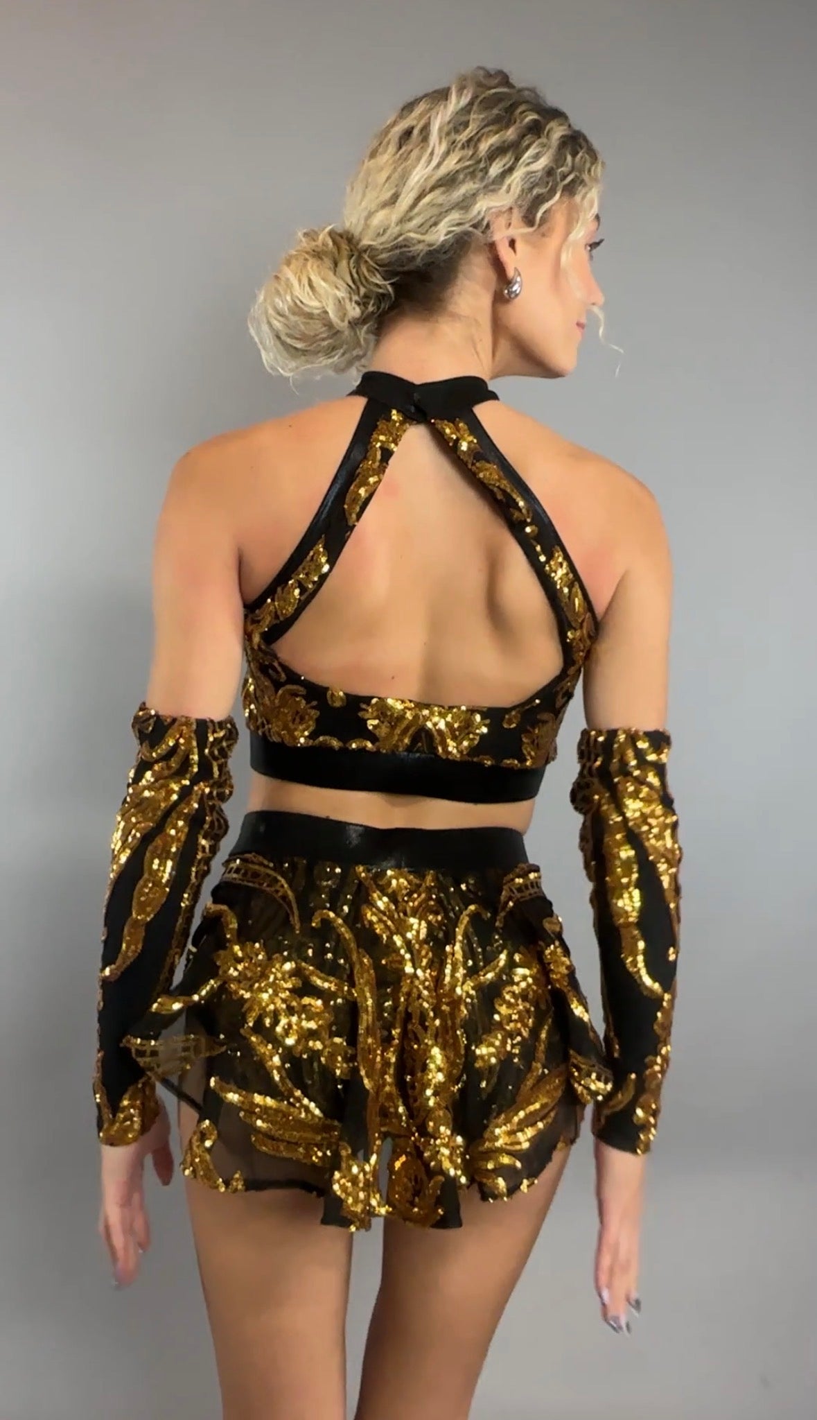 A woman is seen from the back against a gray background, wearing the Gold Sequin on Black Crop Top and Bottom Set featuring a halterneck top with golden sequins, black mesh accents, and coordinating hand accessories.
