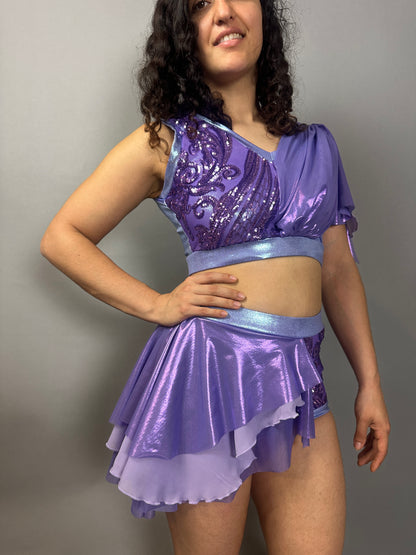 A woman wears a Lavender Purple Sequin Dance Costume featuring high waist shorts, a half skirt, and a crop top with 4-way stretch mesh, posing against a plain background.