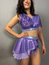 A woman wears a Lavender Purple Sequin Dance Costume featuring high waist shorts, a half skirt, and a crop top with 4-way stretch mesh, posing against a plain background.